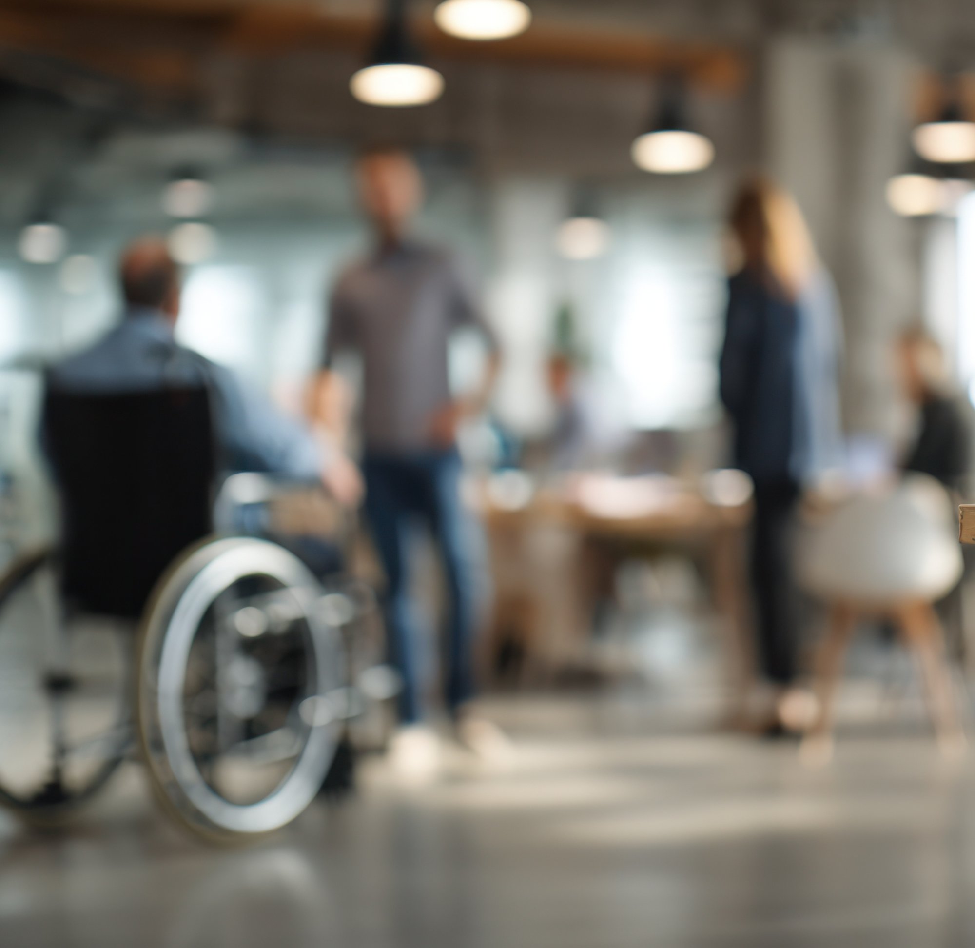 A blurred image of an office setting with a few people talking to a person in a wheelchair. Two of them are standing, and the other three are sitting at their desks. The atmosphere is relaxed, and the colors are warm and subtle.
